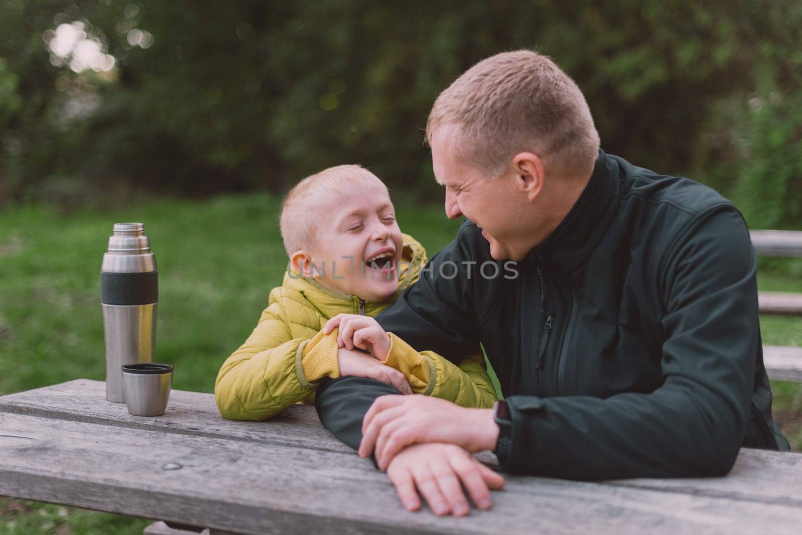 Happy family: father and child boy son playing and laughing in autumn park, sitting on wooden bench. Father and little kid having fun outdoors, playing together. Father and son sitting on a bench and talking. dad son park bench table autumn thermos.