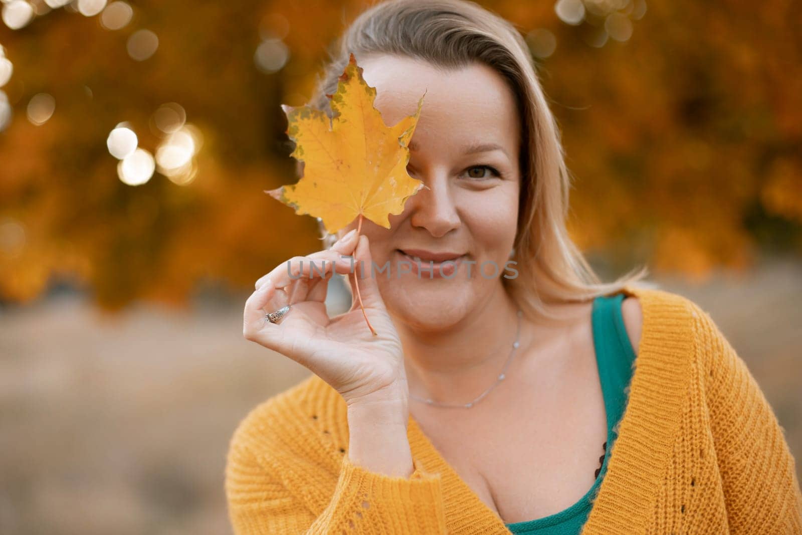 an autumn woman in a green dress holds an autumn leaf in front of her face, against the background of an autumn tree.
