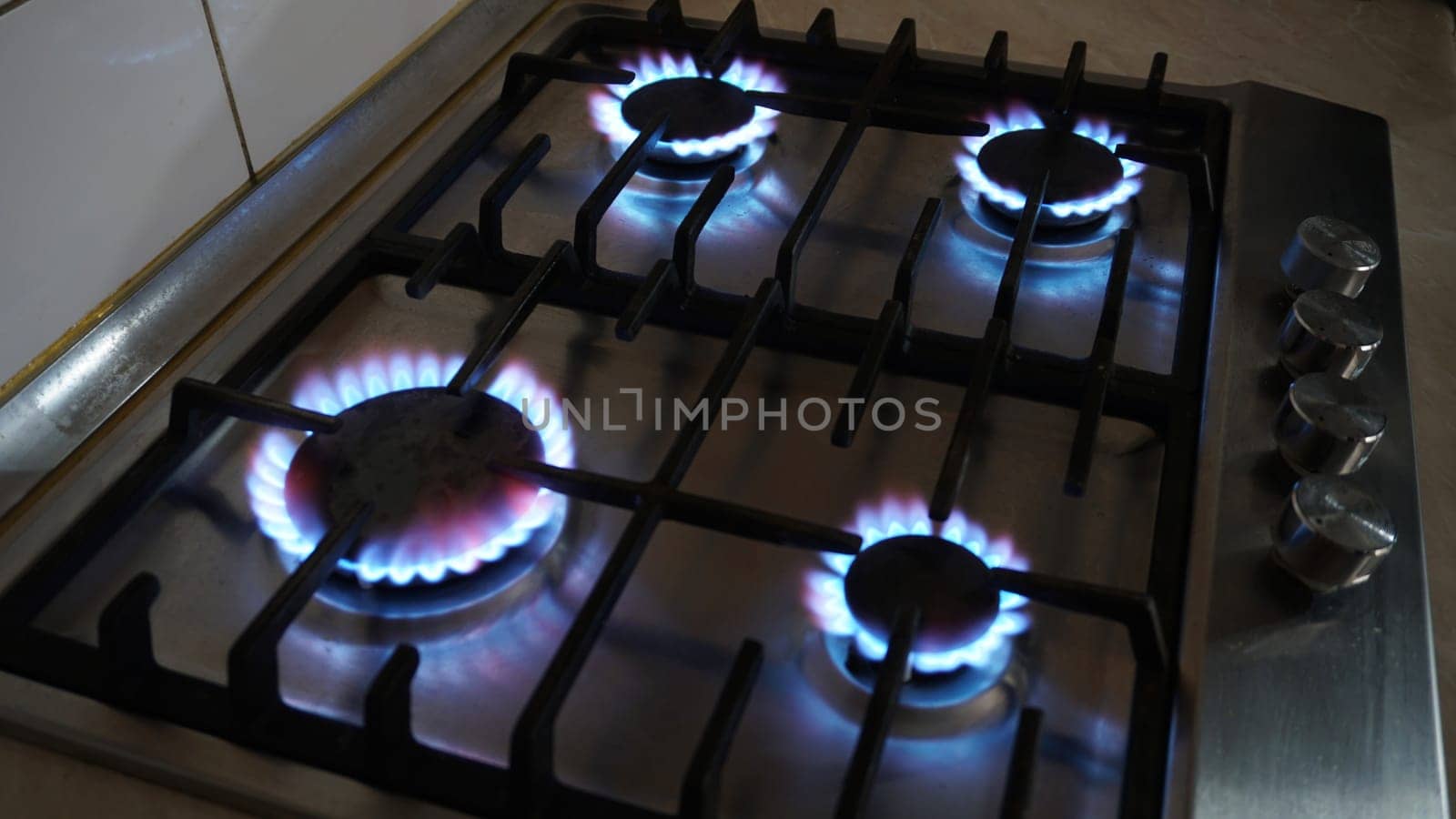 A blue fire is burning on the gas stove. Gas at home. Metal gas stove. Black bars. Light switches of burners. The gas is working. Beige tiles on the walls. Stop the economic crisis. Warmth in house