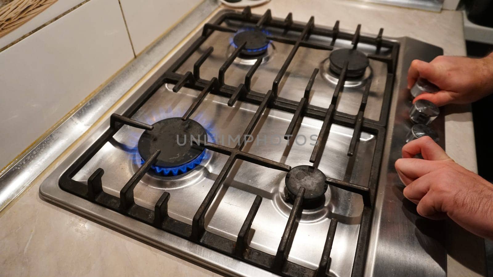 Turns off or turns on the gas on the stove. Blue flame of fire on black burners. The man's hands turn off or turn on the gas. Black grilles on a steel plate. White tiles on the walls. Cold winter
