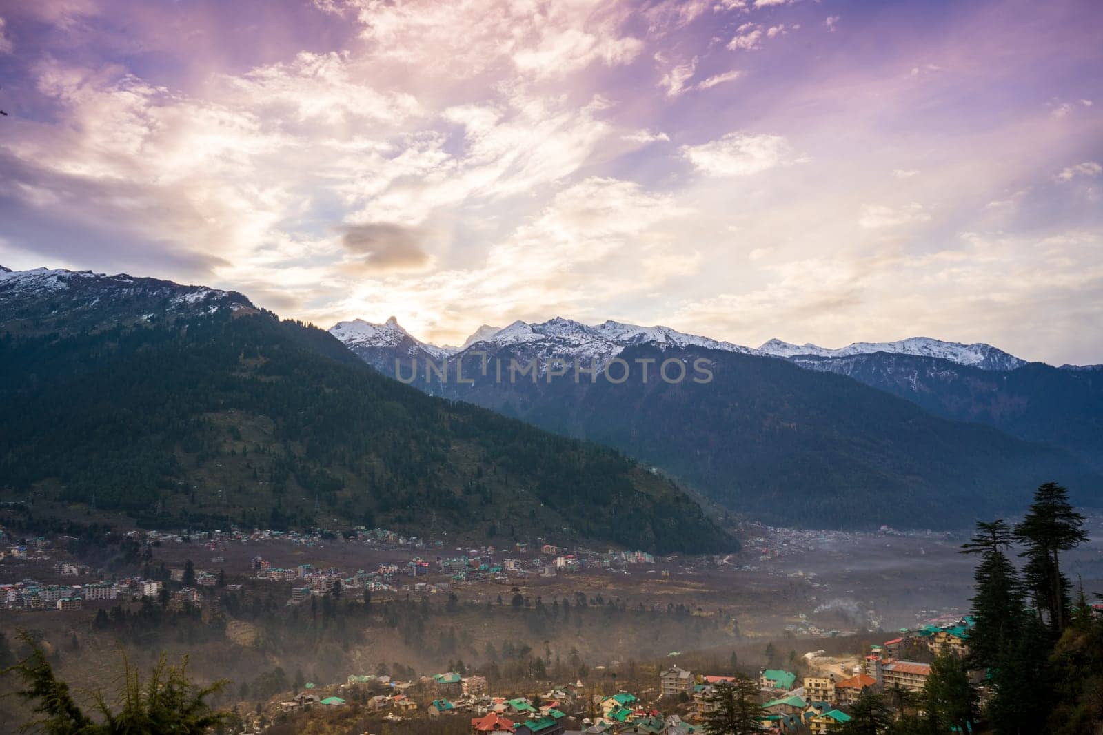 monsoon clouds moving over snow covered himalaya mountains with the blue orange sunset sunrise light with town of kullu manali valley at the base of mountains by Shalinimathur