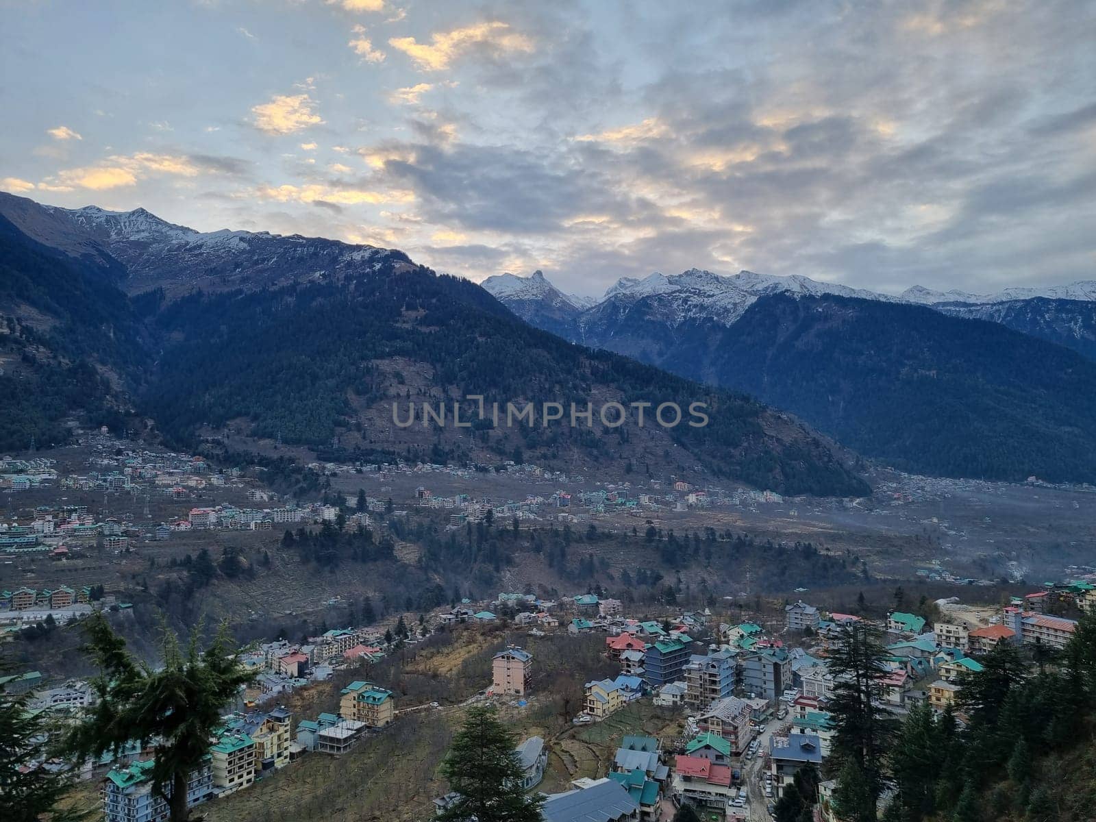 monsoon clouds moving over snow covered himalaya mountains with the blue orange sunset sunrise light with town of kullu manali valley at the base of mountains India