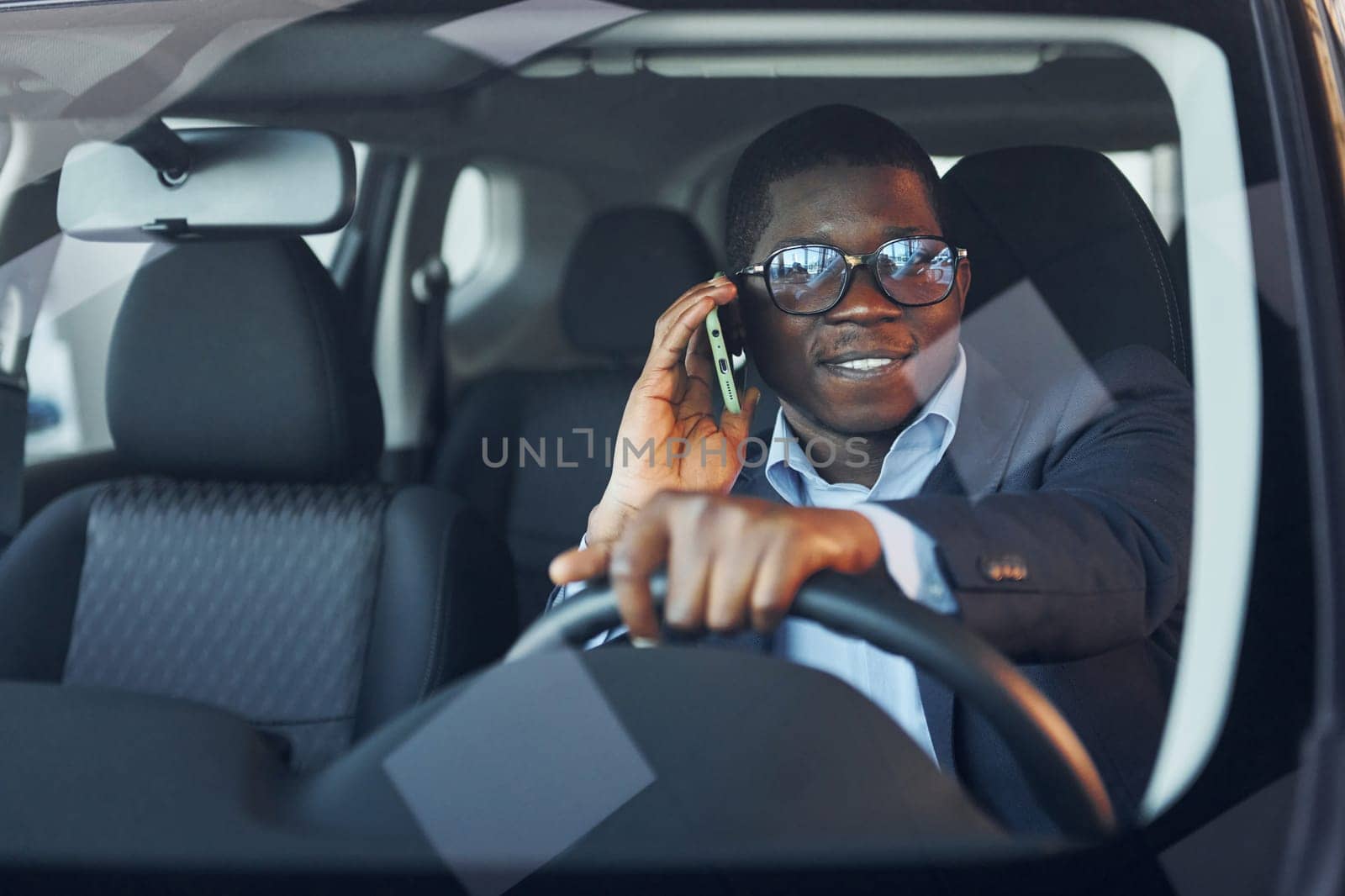 Front view. Young african american businessman in black suit is in the automobile.