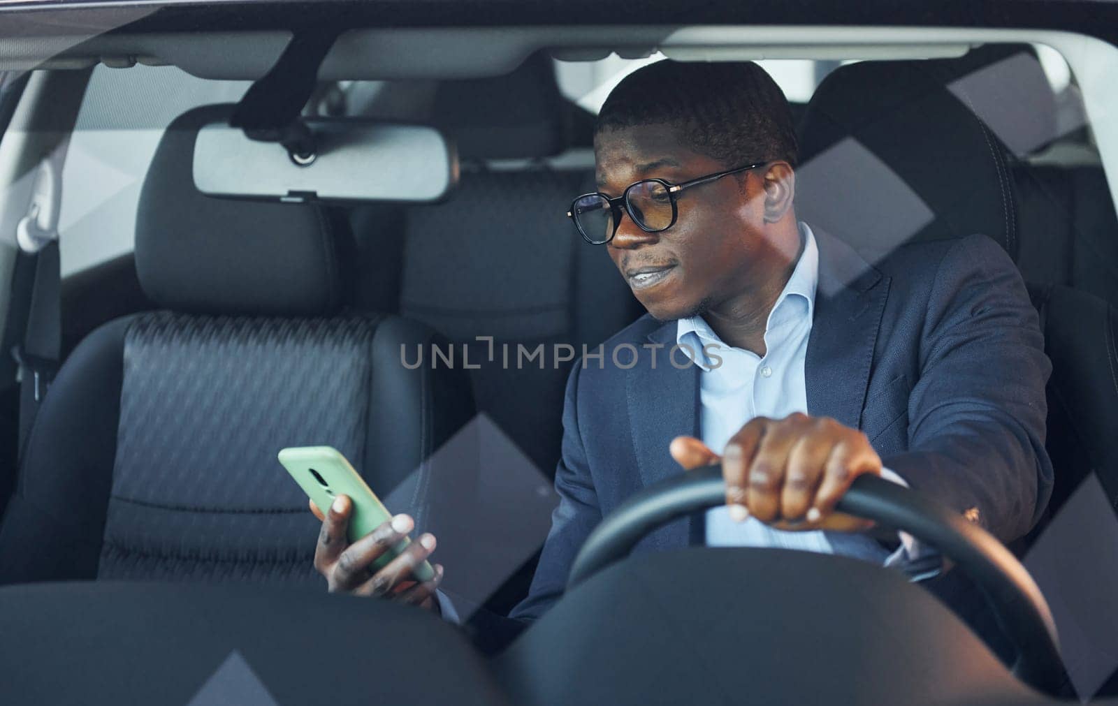 With phone. Young african american businessman in black suit is in the automobile.