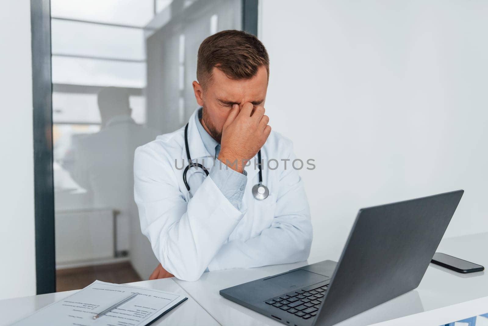 Using laptop and feeling tired. Professional medic in white coat is in the clinic.