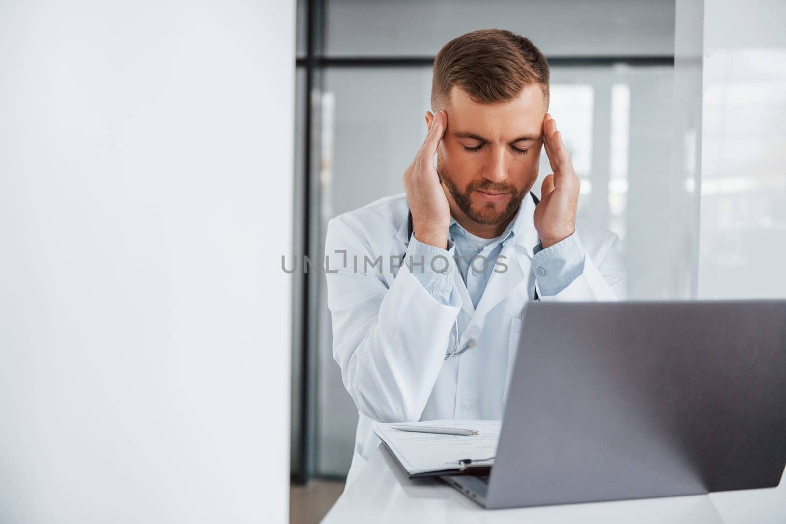 Using laptop and feeling tired. Professional medic in white coat is in the clinic.