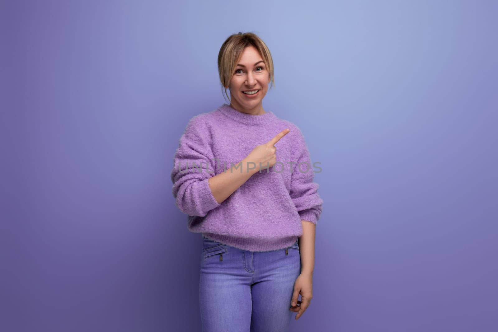 smiling cute blond young woman in a lilac sweater points a finger to the side on a bright background with copy space by TRMK