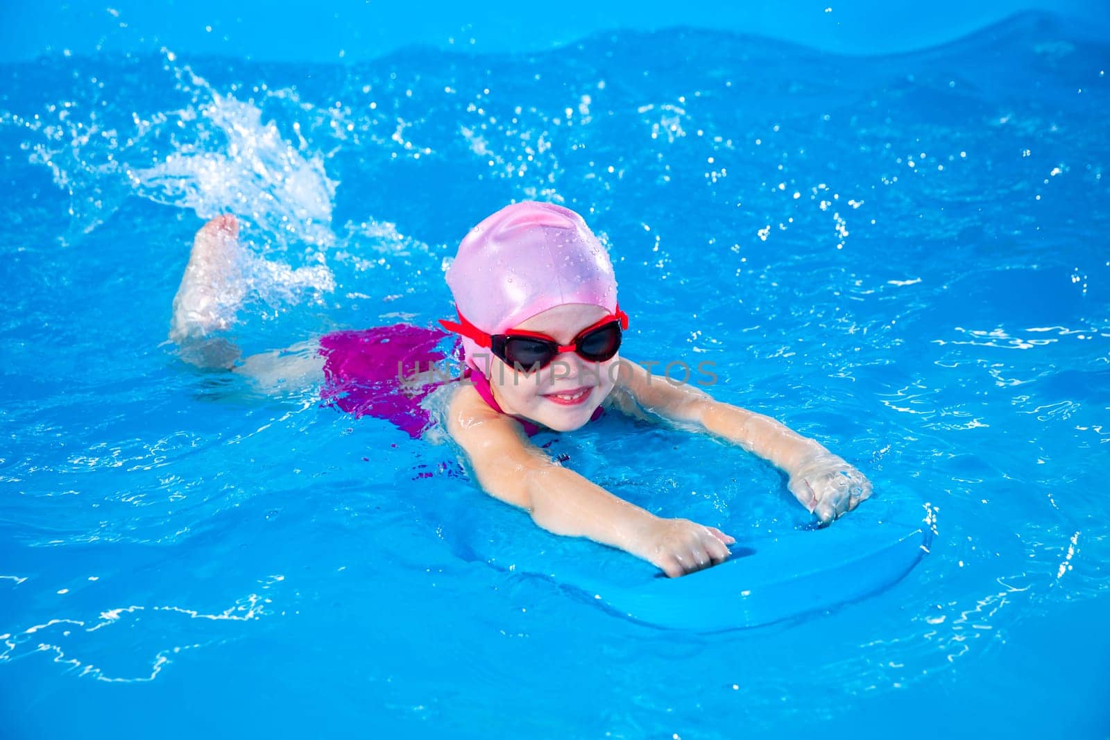 Smiling little girl learning to swim in indoor pool with flutter board during swimming class