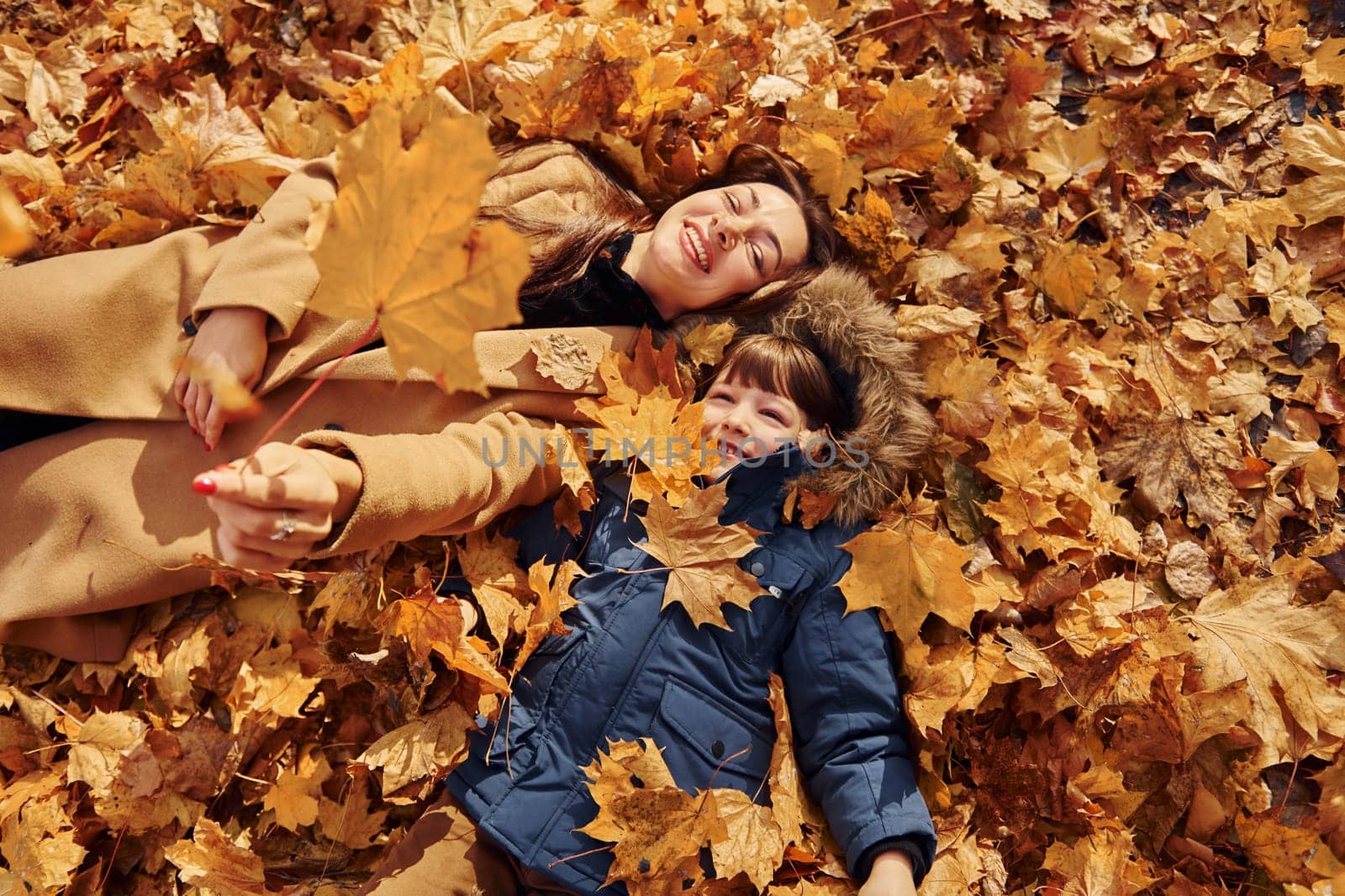 Laying down on the leaves. Mother with her son is having fun outdoors in the autumn forest by Standret
