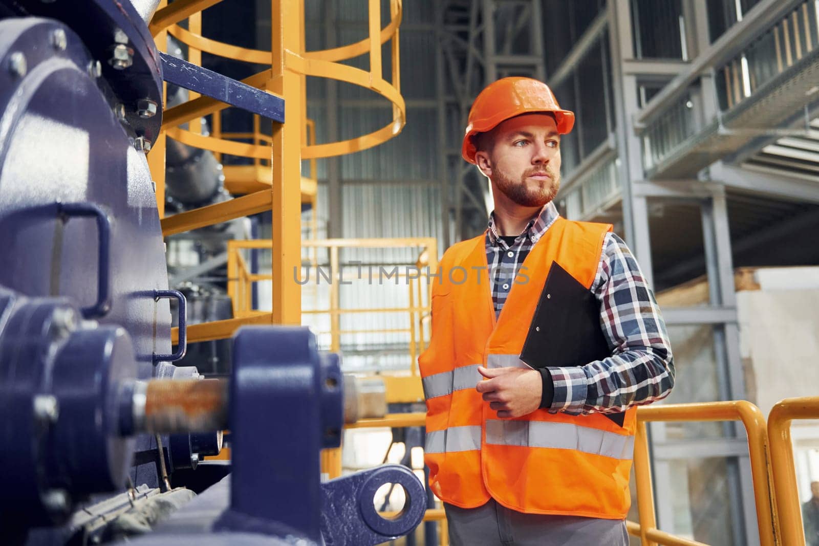 With notepad. Construction worker in uniform is in the factory.