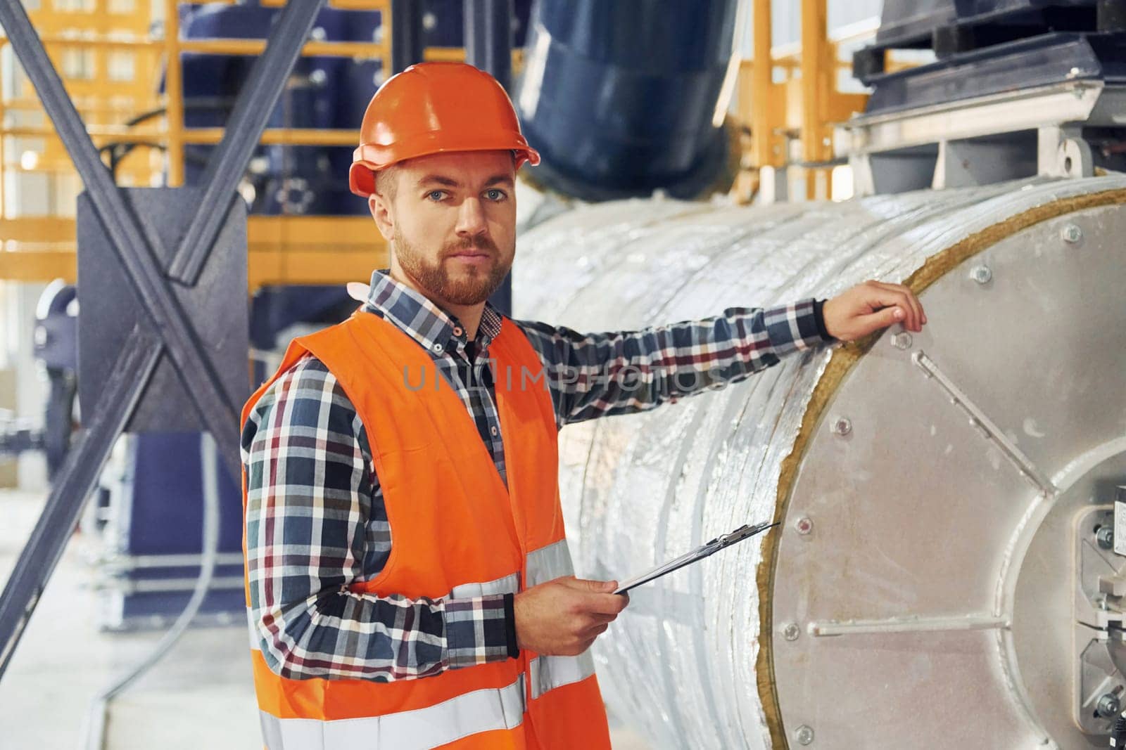 In orange colored hard hat. Construction worker in uniform is in the factory.