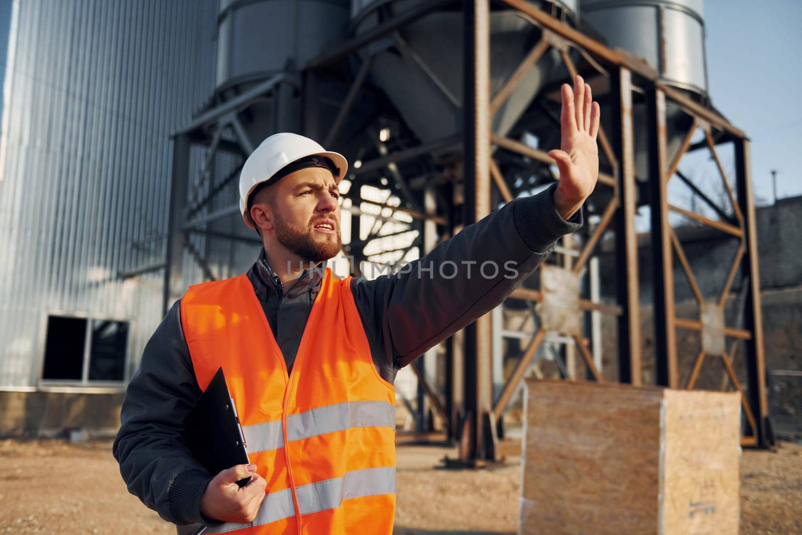 Showing stop gesture. Construction worker in uniform is outdoors near the factory.