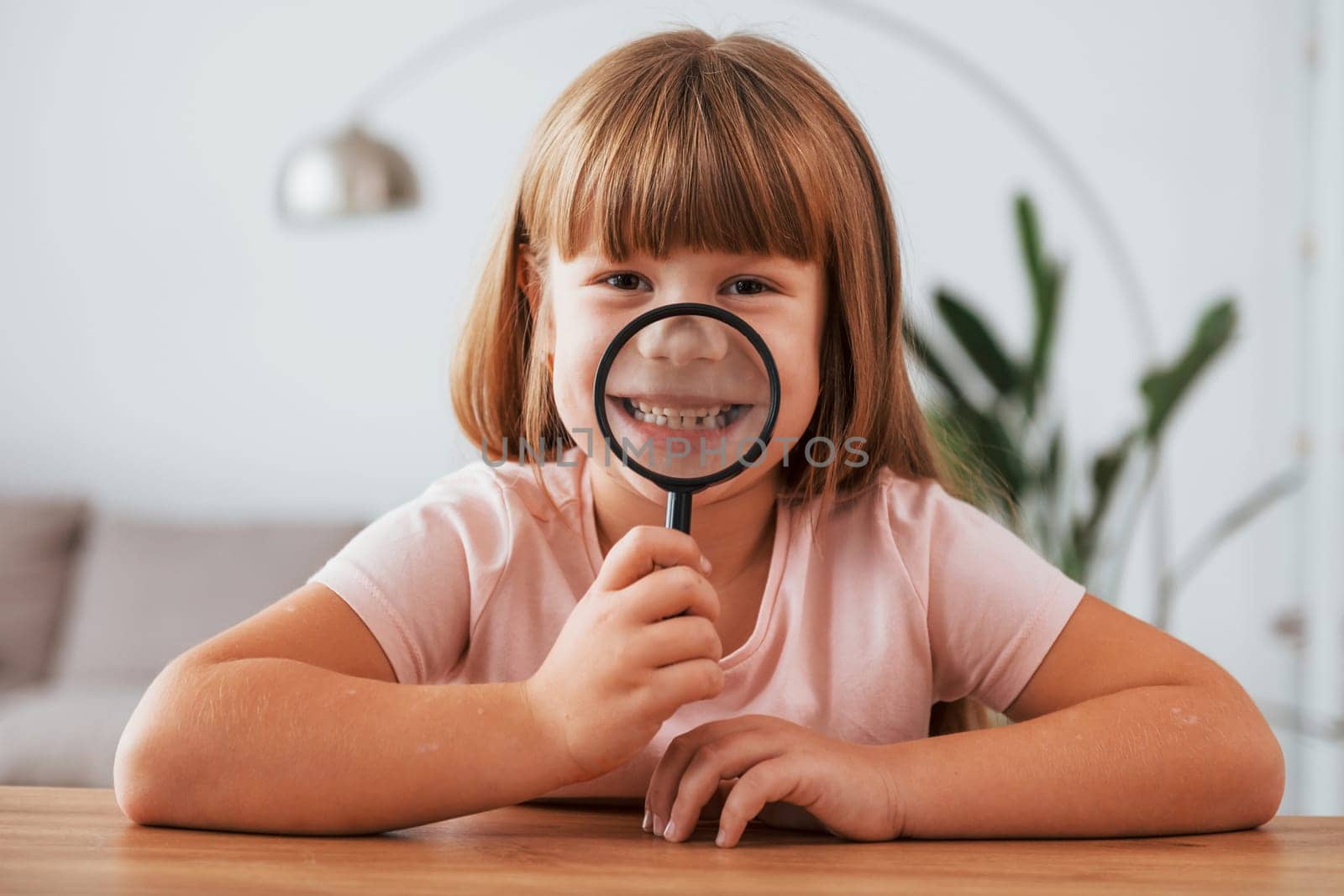 Having fun. Little girl is sitting by the table with magnifying glass.