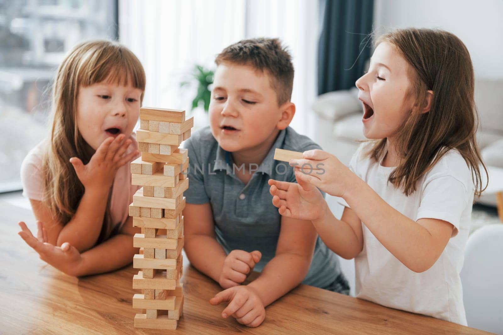 Focused on the jumbling tower game. Kids having fun in the domestic room at daytime together.