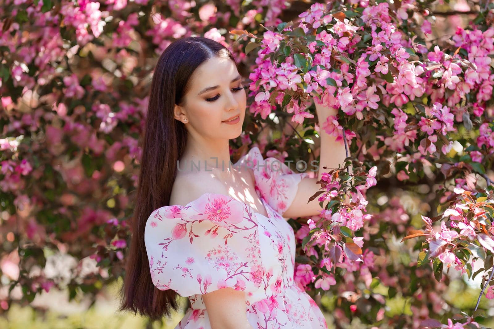 A pretty brunette girl in a pink dress standing near pink blooming apple trees, in the spring in the garden. Side view. Close up. Copy space