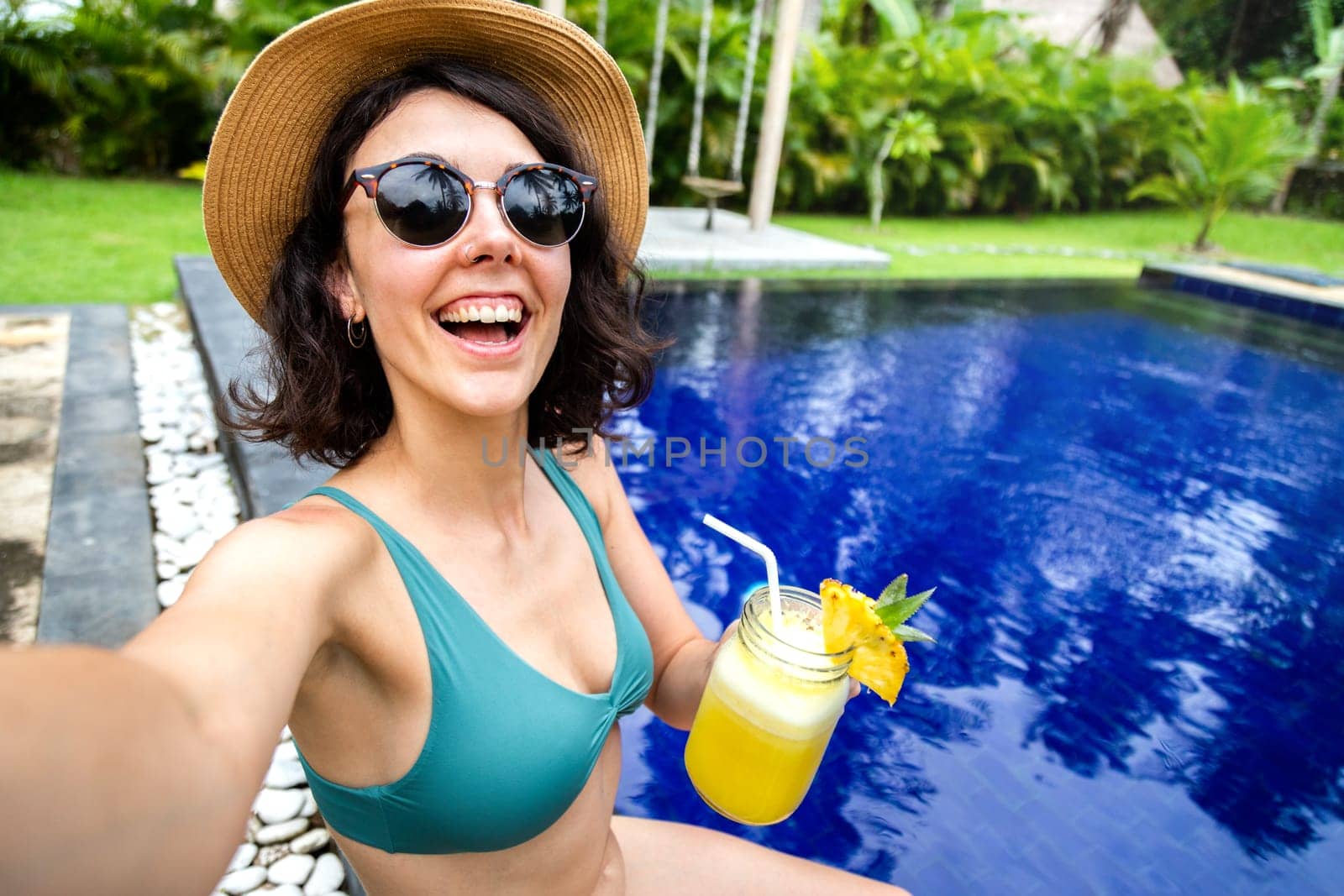 Happy young woman wearing a bikini drinking juice taking selfie in the swimming pool during summer vacation in tropical resort. Holiday concept.