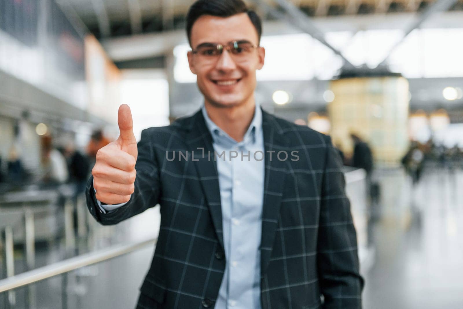 Showing thumb up. Young businessman in formal clothes is in the airport at daytime.