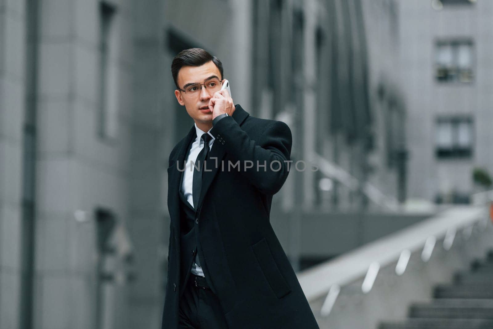 Talking by phone. Businessman in black suit and tie is outdoors in the city.