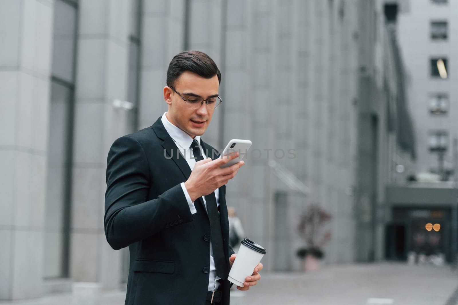 Using phone. Businessman in black suit and tie is outdoors in the city.