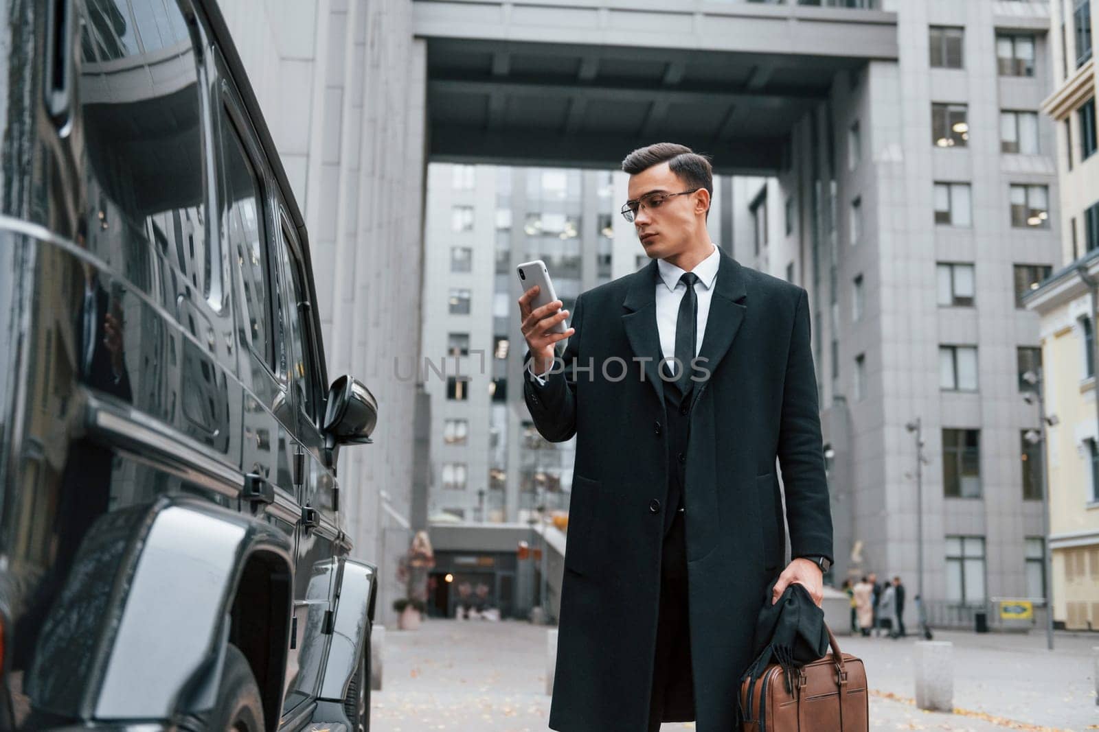 Standing near black car. Businessman in black suit and tie is outdoors in the city.