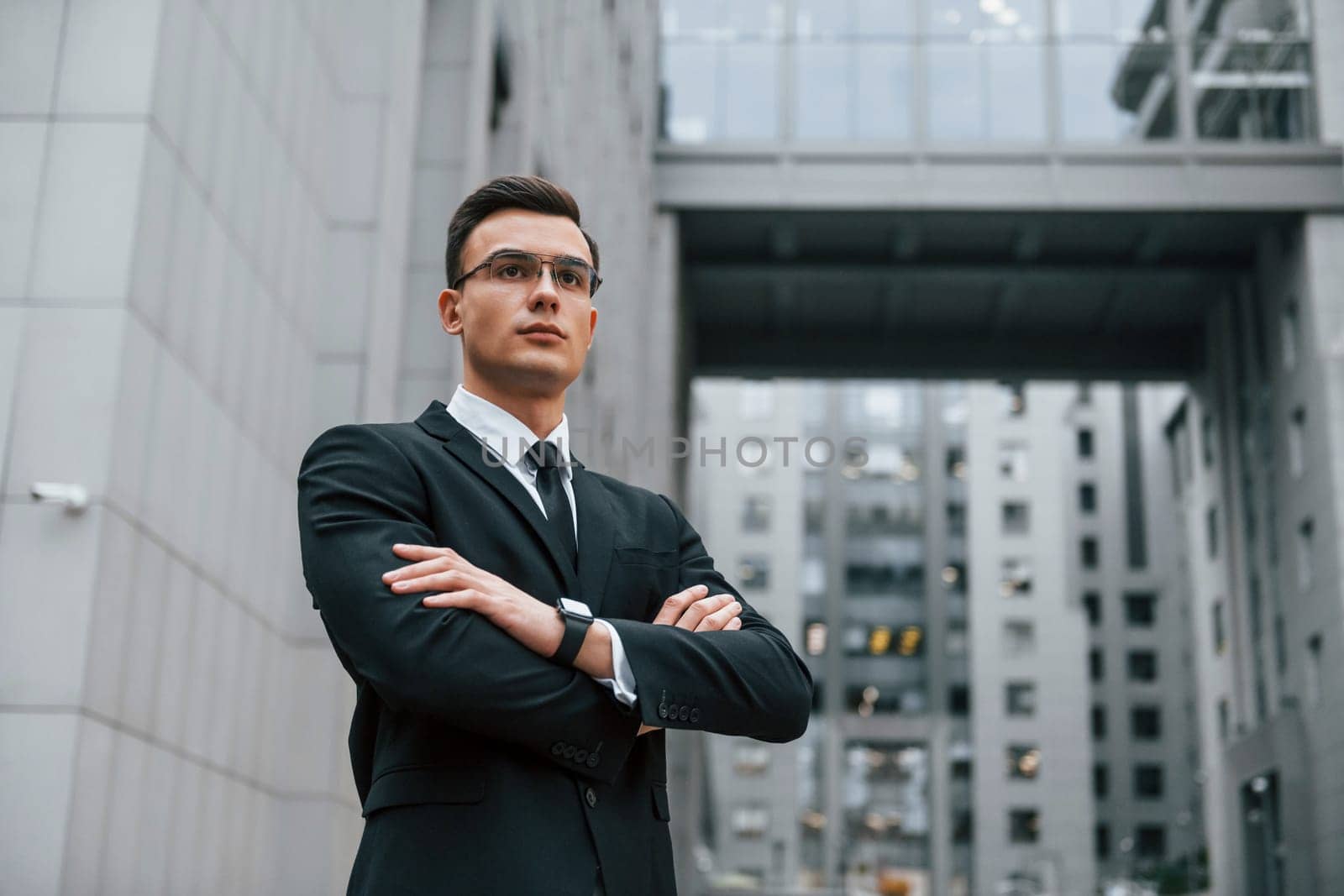 Standing with arms crossed. Businessman in black suit and tie is outdoors in the city.