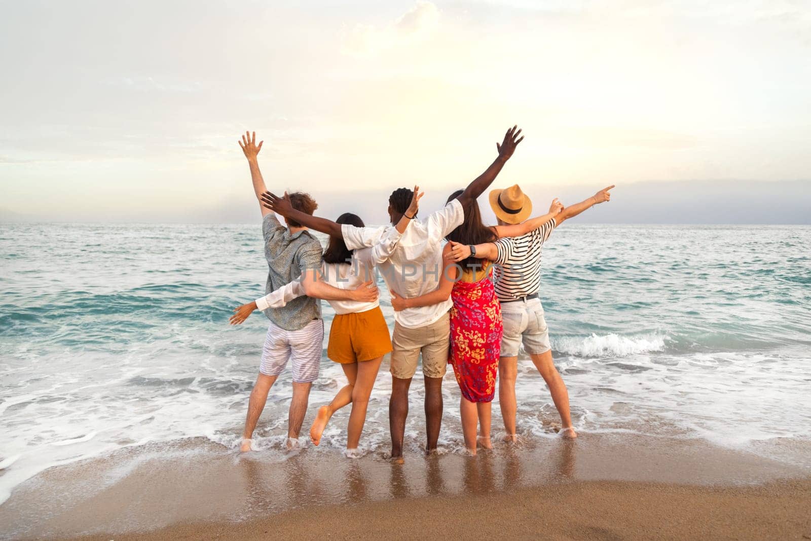 Rear view of multiracial friends embracing together looking at the ocean celebrating with arms up during vacation trip. Friendship concept.
