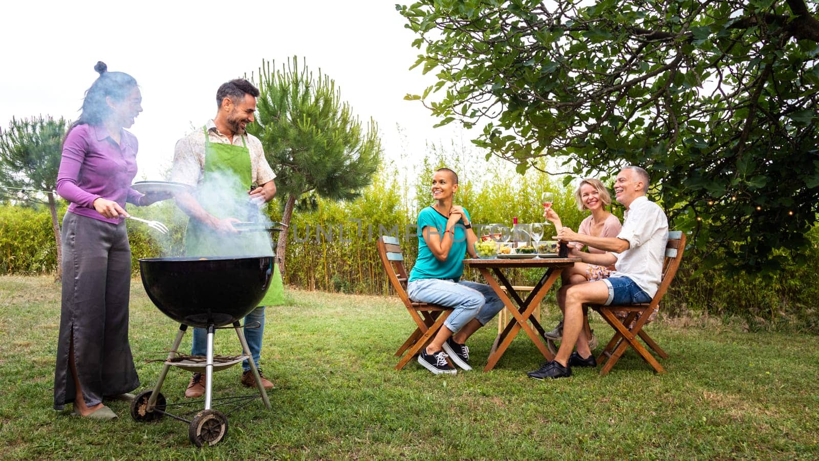 Multiracial couple cooking food on grill for friends. Outdoor garden barbecue party. Friends laughing and having fun, enjoying wine in backyard. Lifestyle.