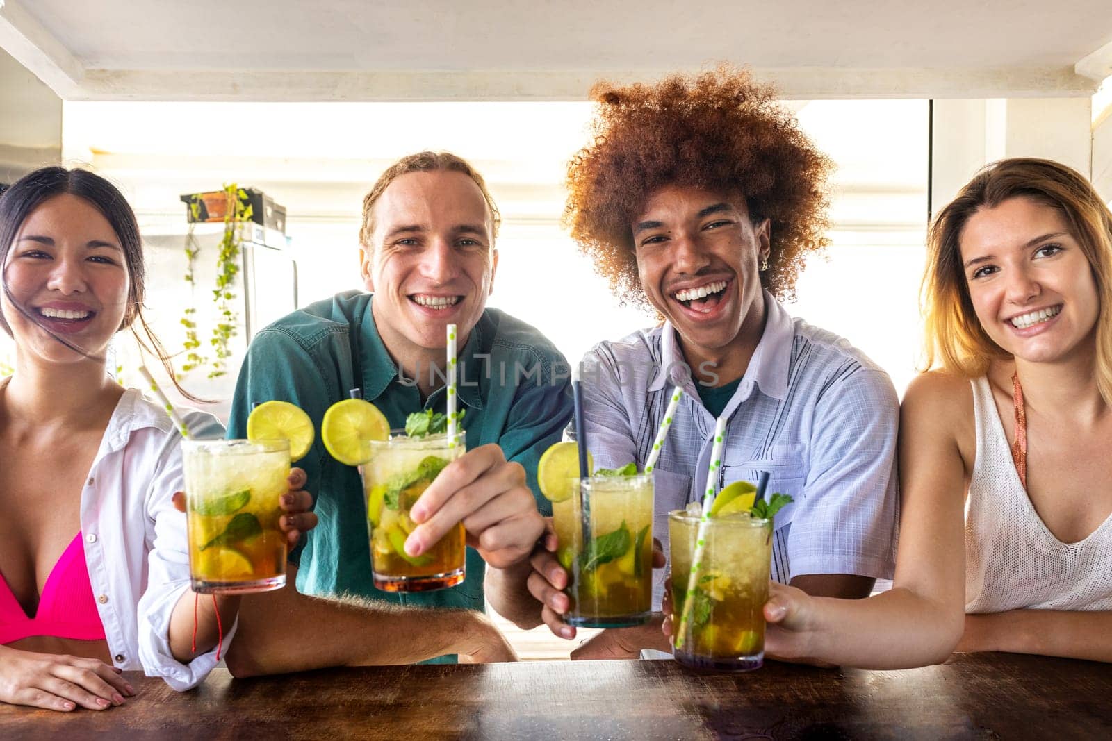 Multiracial friends toasting with cocktails at beach bar looking at camera. Happy friends having fun at summertime. by Hoverstock