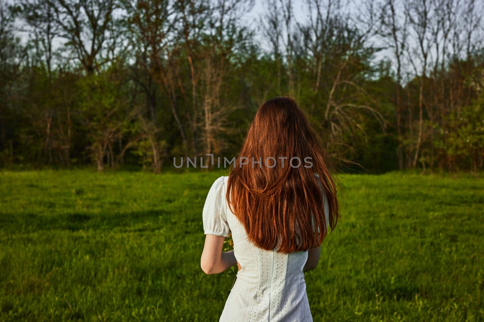 a woman with long red hair in a white dress stands in a green field at sunset with her back to the camera. High quality photo