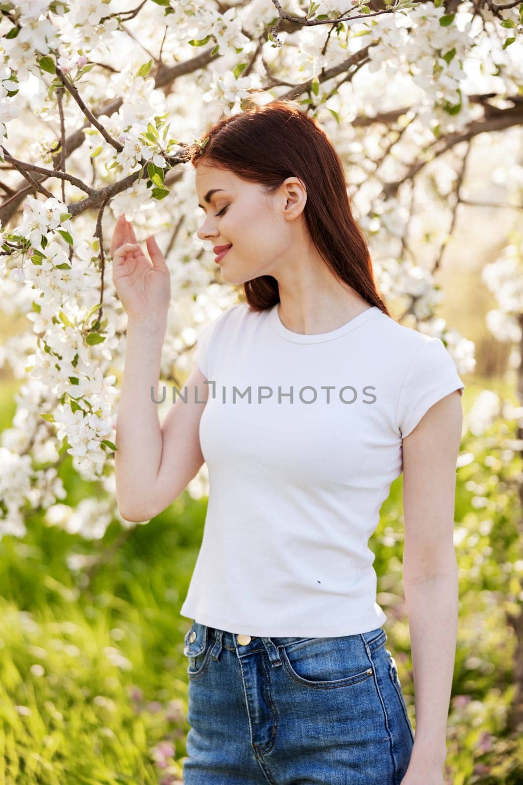 portrait of a happy woman with red hair in casual clothes touching flowers on the branches of a fruit tree by Vichizh