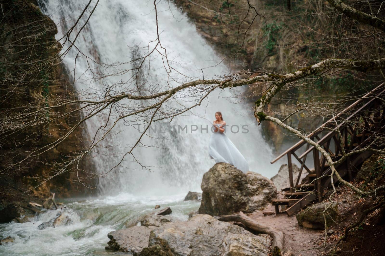 Happy woman in a white dress stands on a stone with a waterfall behind.