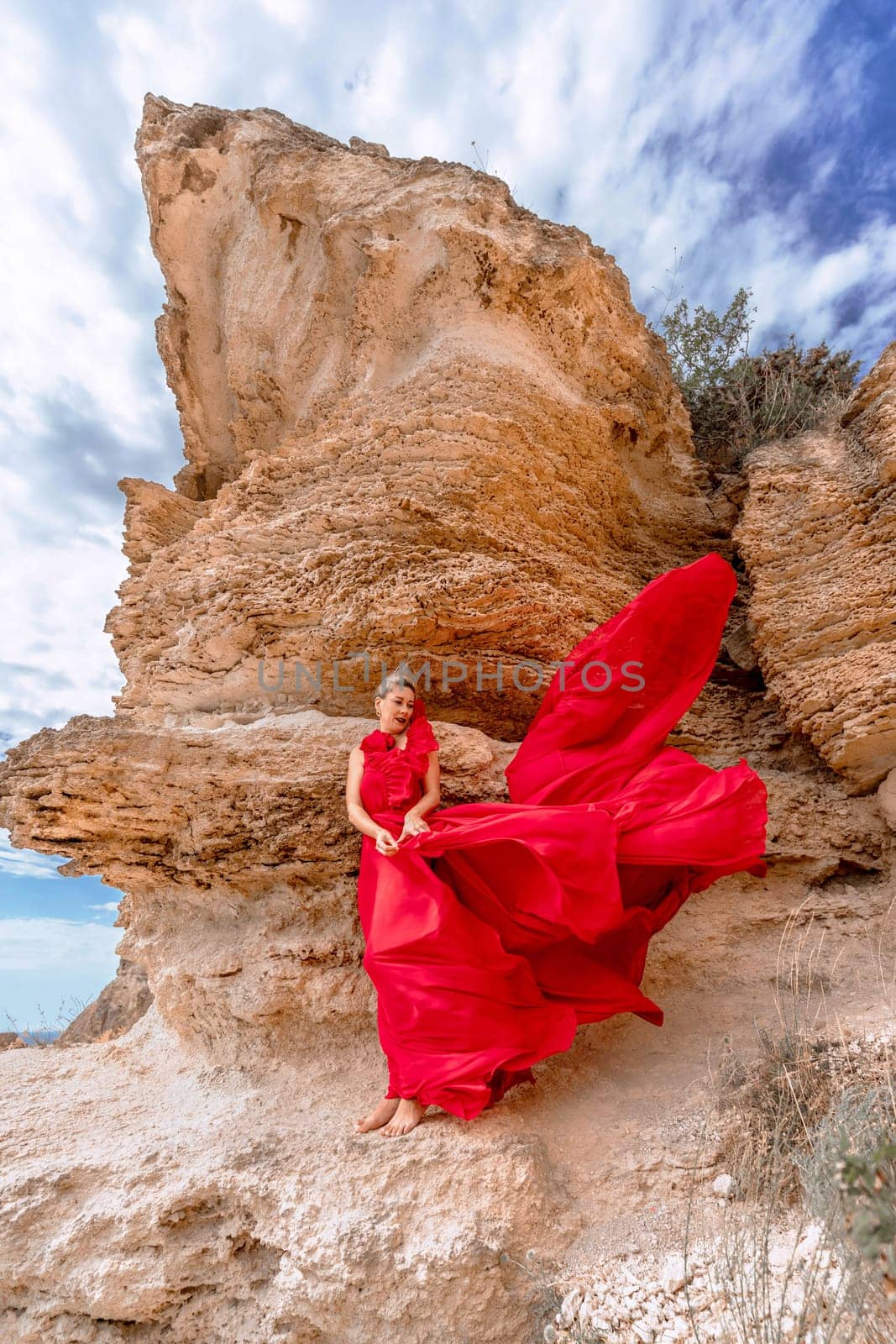 A woman in a red silk dress stands by the ocean, with mountains in the background, as her dress sways in the breeze