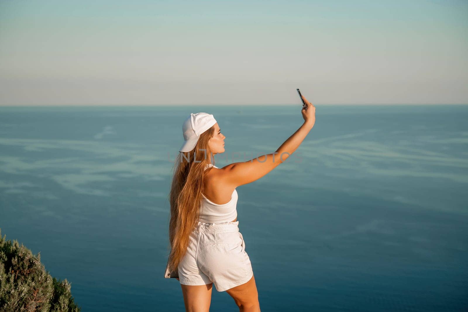 Selfie woman in a cap, white tank top and shorts makes a selfie shot mobile phone post photo social network outdoors on the background of the sea beach people vacation lifestyle travel concept. by Matiunina
