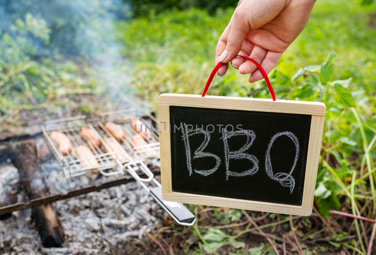 Close-up of fried sausages on the grill. A man is holding a sign with the inscription barbecue. Rest in nature, picnic, weekend. by sfinks