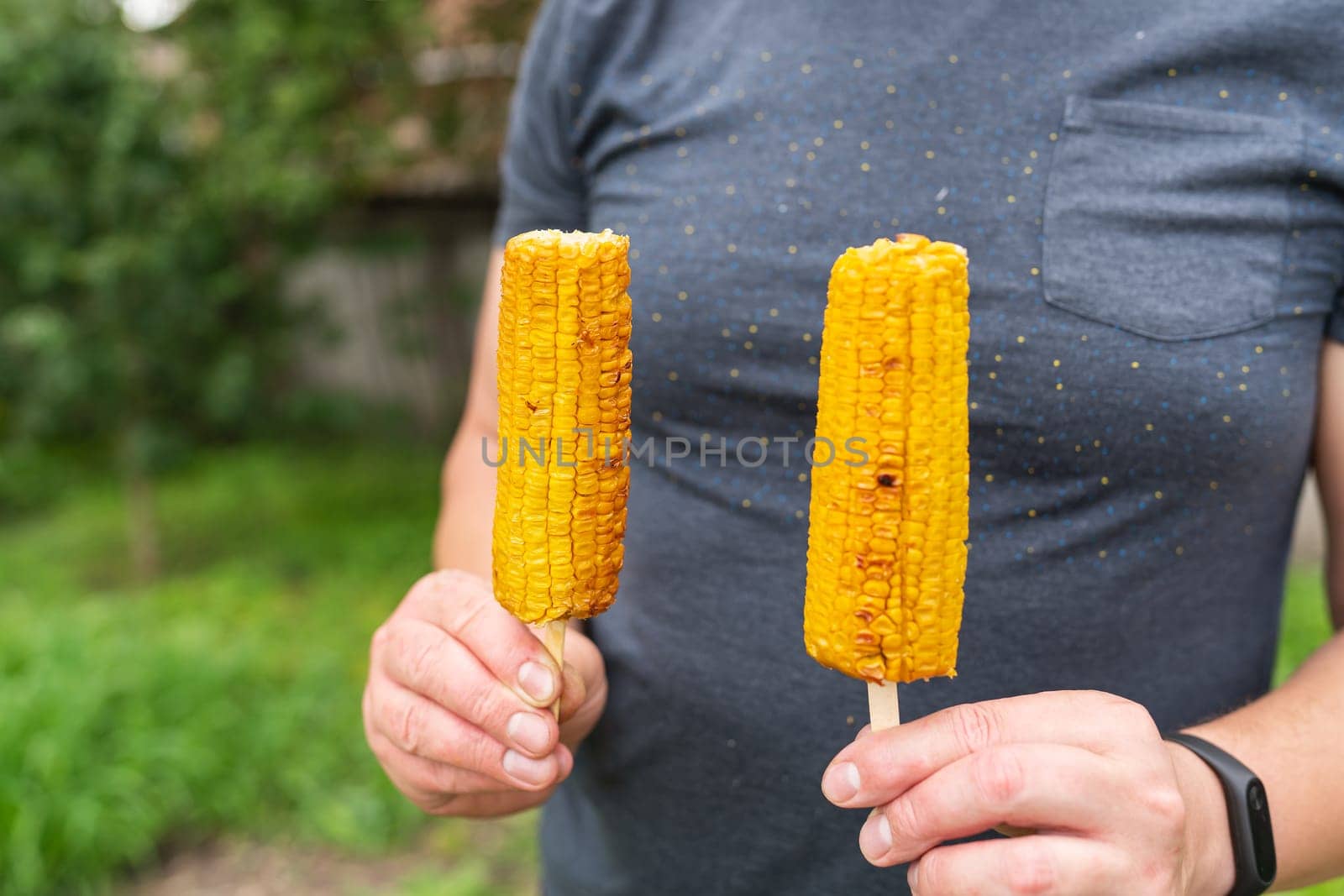 In his hands, a man holds roasted sweet corn with salt and butter on a stick on a grill on the background of a recreation park. Summer vegan dinner or appetizer, grilling