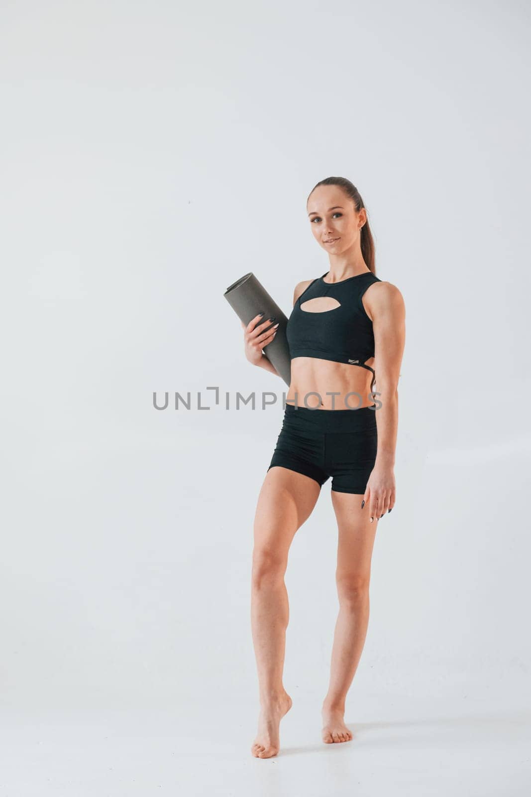 Holding yoga mat and standing. Young woman in sportive clothes doing gymnastics indoors.