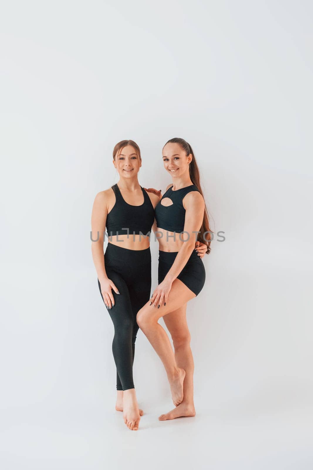 Two young women in sportive clothes is together indoors against white background.