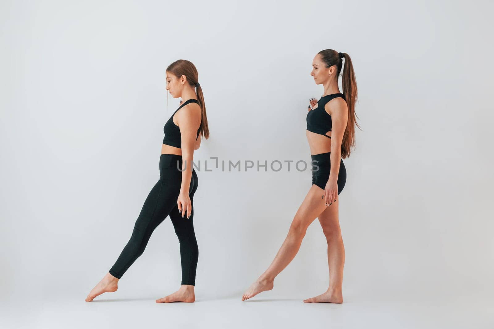 Two young women in sportive clothes is together indoors against white background.