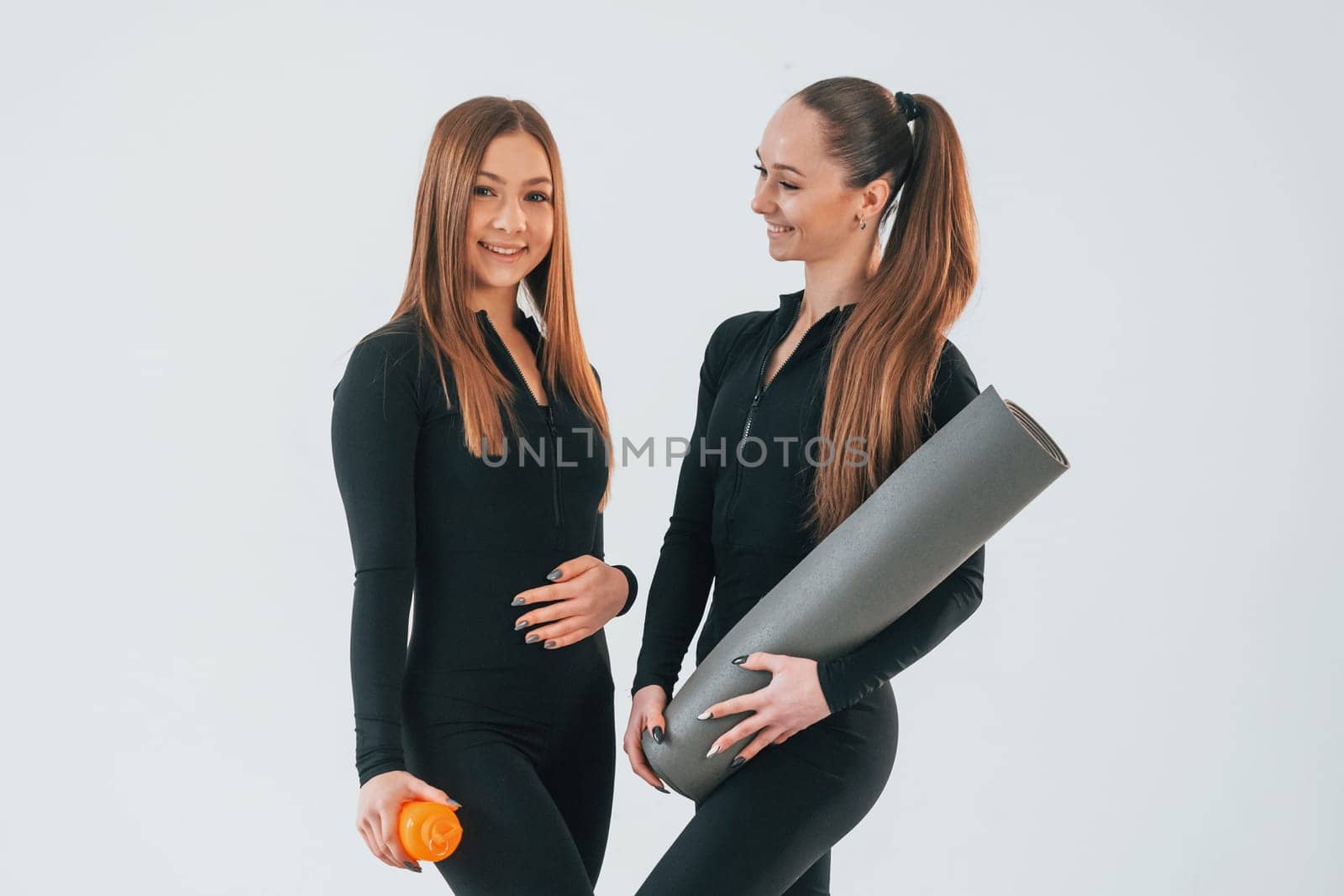 Holding mat. Two young women in sportive clothes is together indoors against white background.