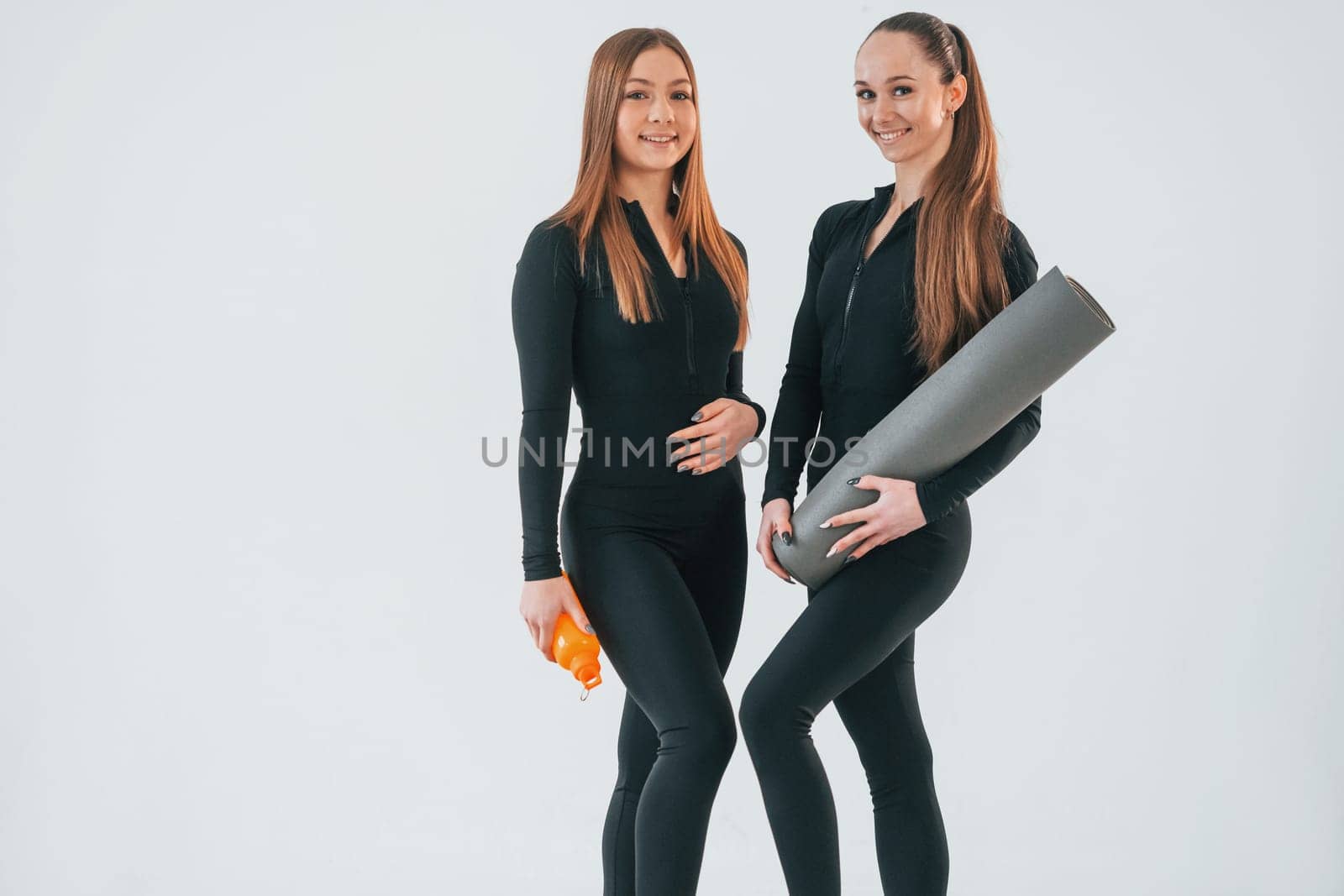 Holding mat. Two young women in sportive clothes is together indoors against white background.