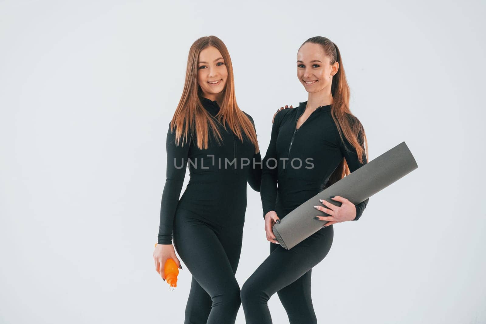 Holding mat. Two young women in sportive clothes is together indoors against white background.