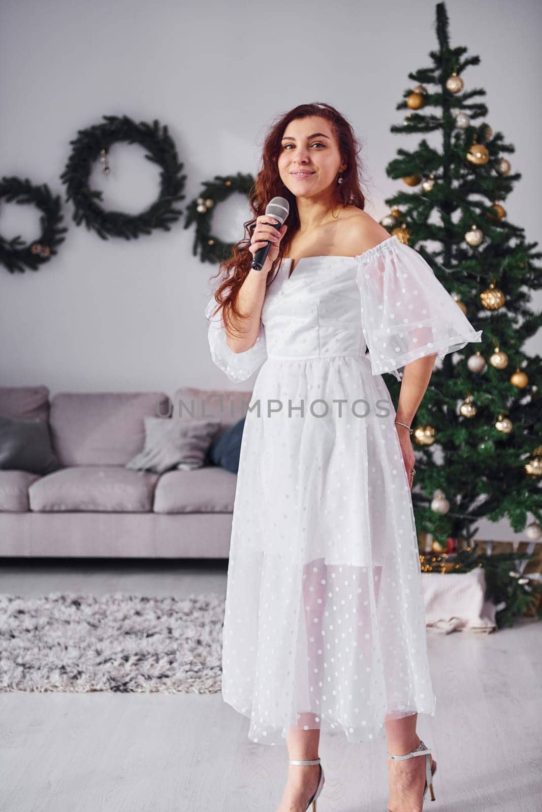 Woman in white dress and with microphone in hands is singing in the christmas decorated domestic room.