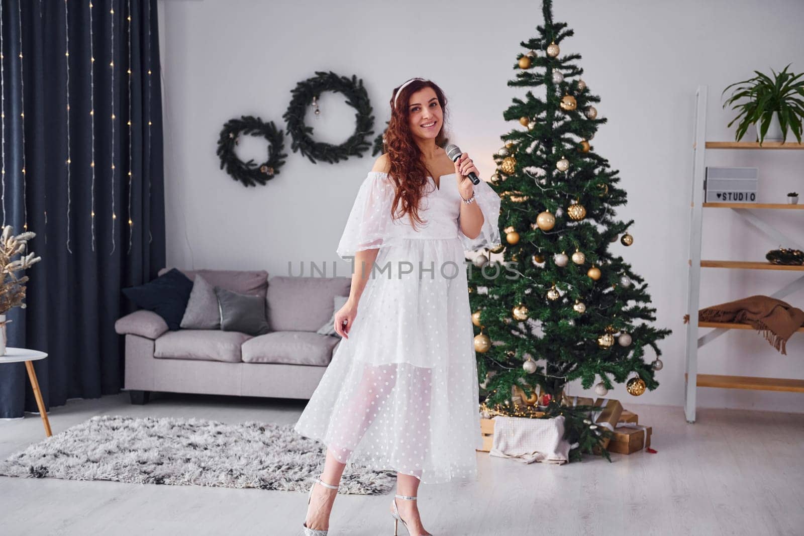 Woman in white dress and with microphone in hands is singing in the christmas decorated domestic room.