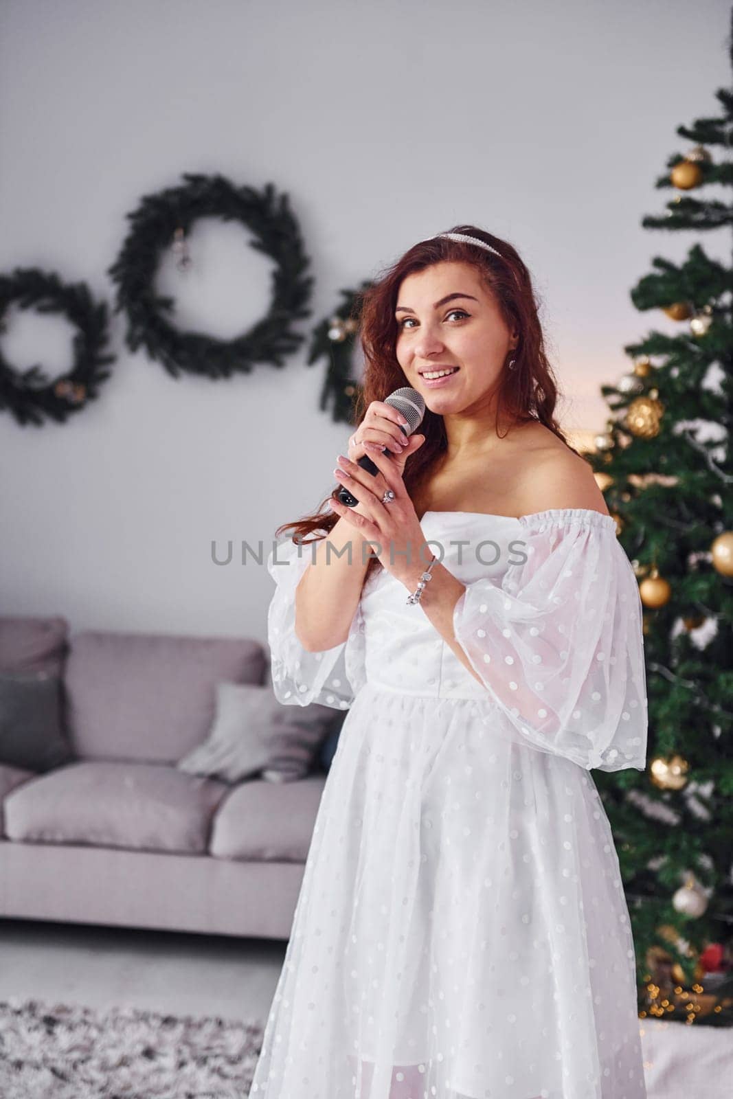 Standing in the christmas decorated domestic room. Woman in white dress and with microphone in hands is singing in the .