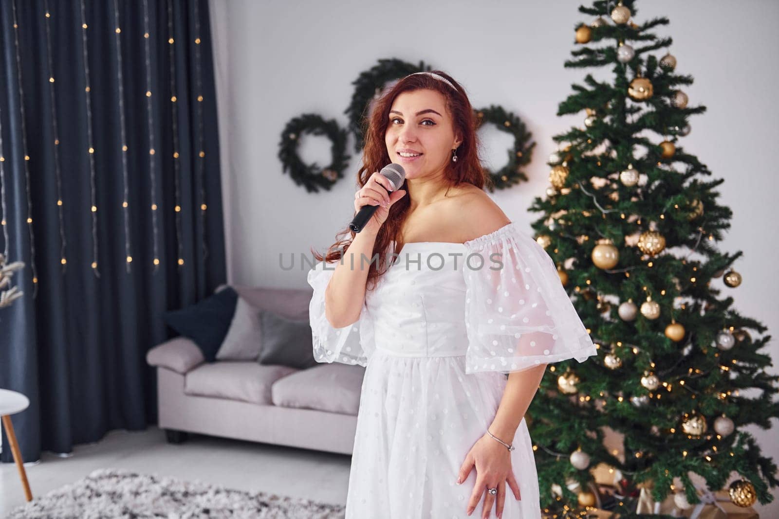 Standing in the christmas decorated domestic room. Woman in white dress and with microphone in hands is singing in the .