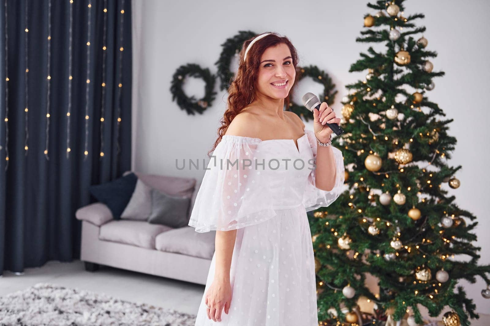 Standing in the christmas decorated domestic room. Woman in white dress and with microphone in hands is singing in the .