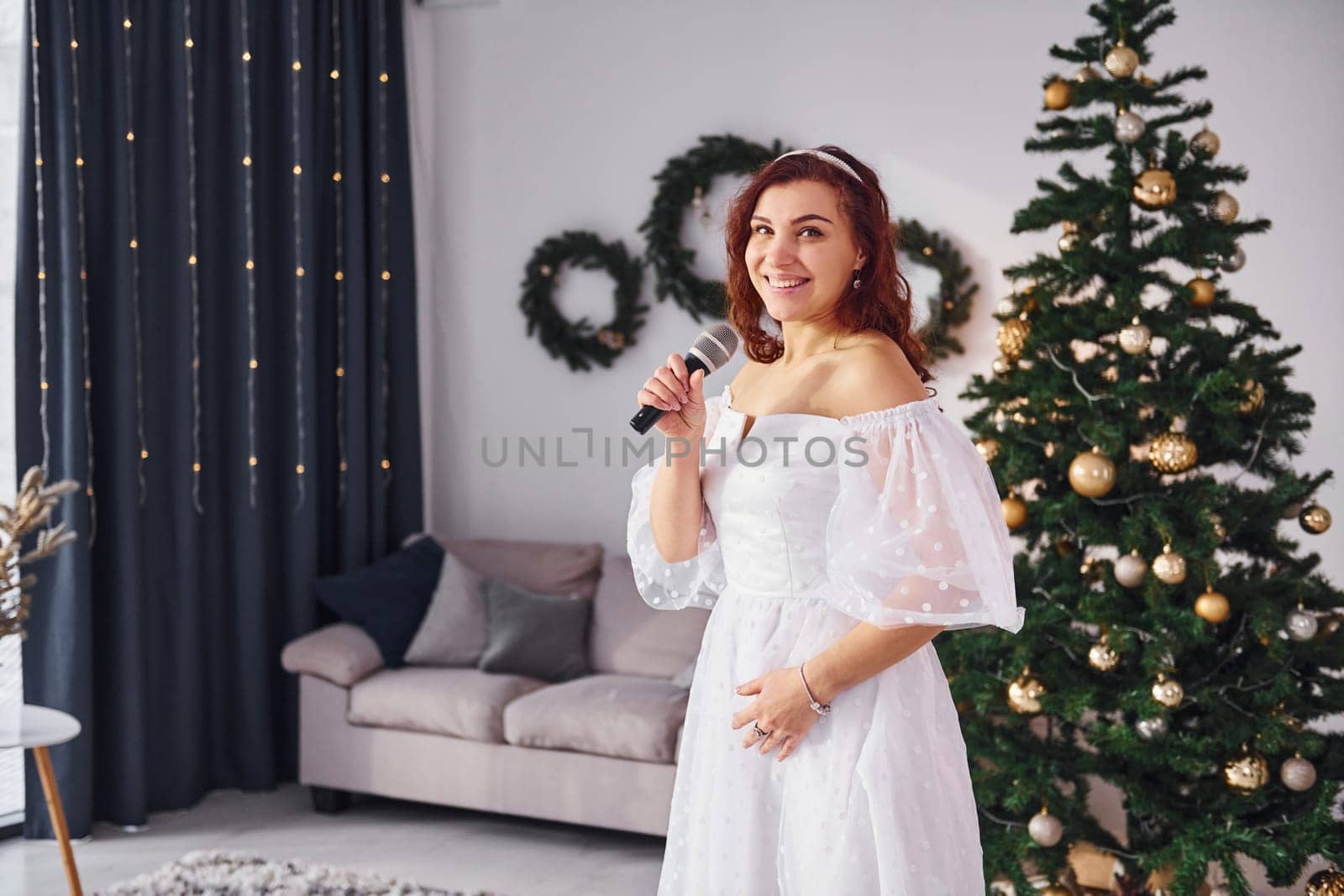 Standing in the christmas decorated domestic room. Woman in white dress and with microphone in hands is singing in the .