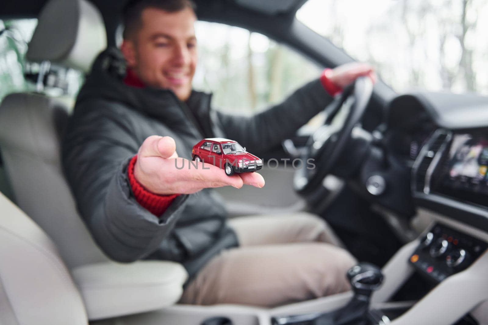 Man sitting in the automobile and holding little red toy car in hand.