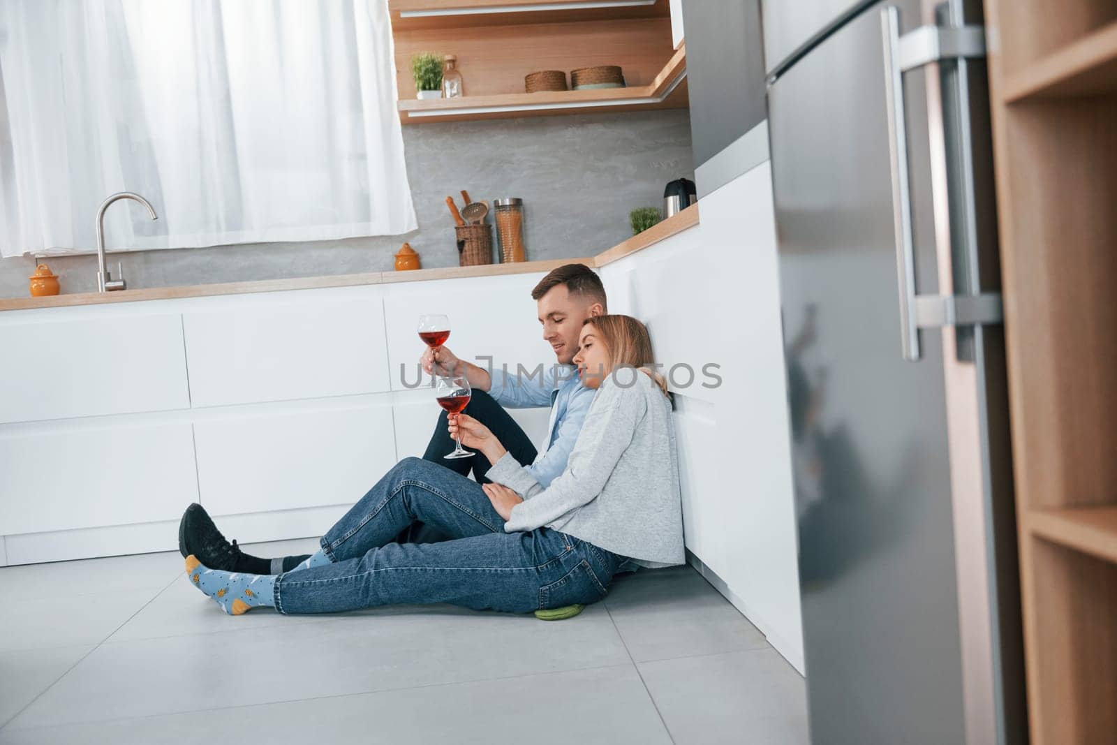 Drinking wine. Couple sitting on the floor of modern kitchen by Standret