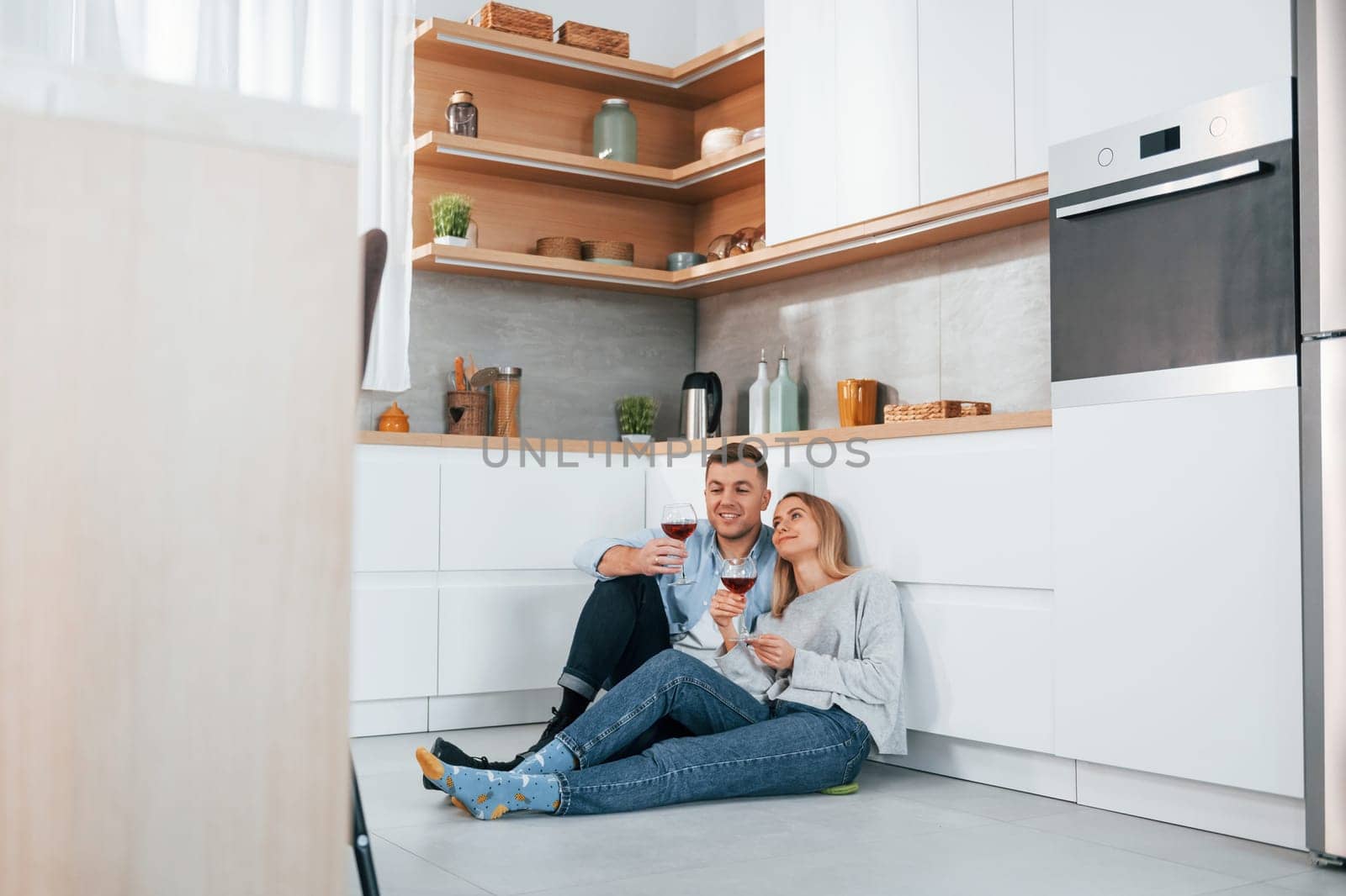 Drinking wine. Couple sitting on the floor of modern kitchen by Standret